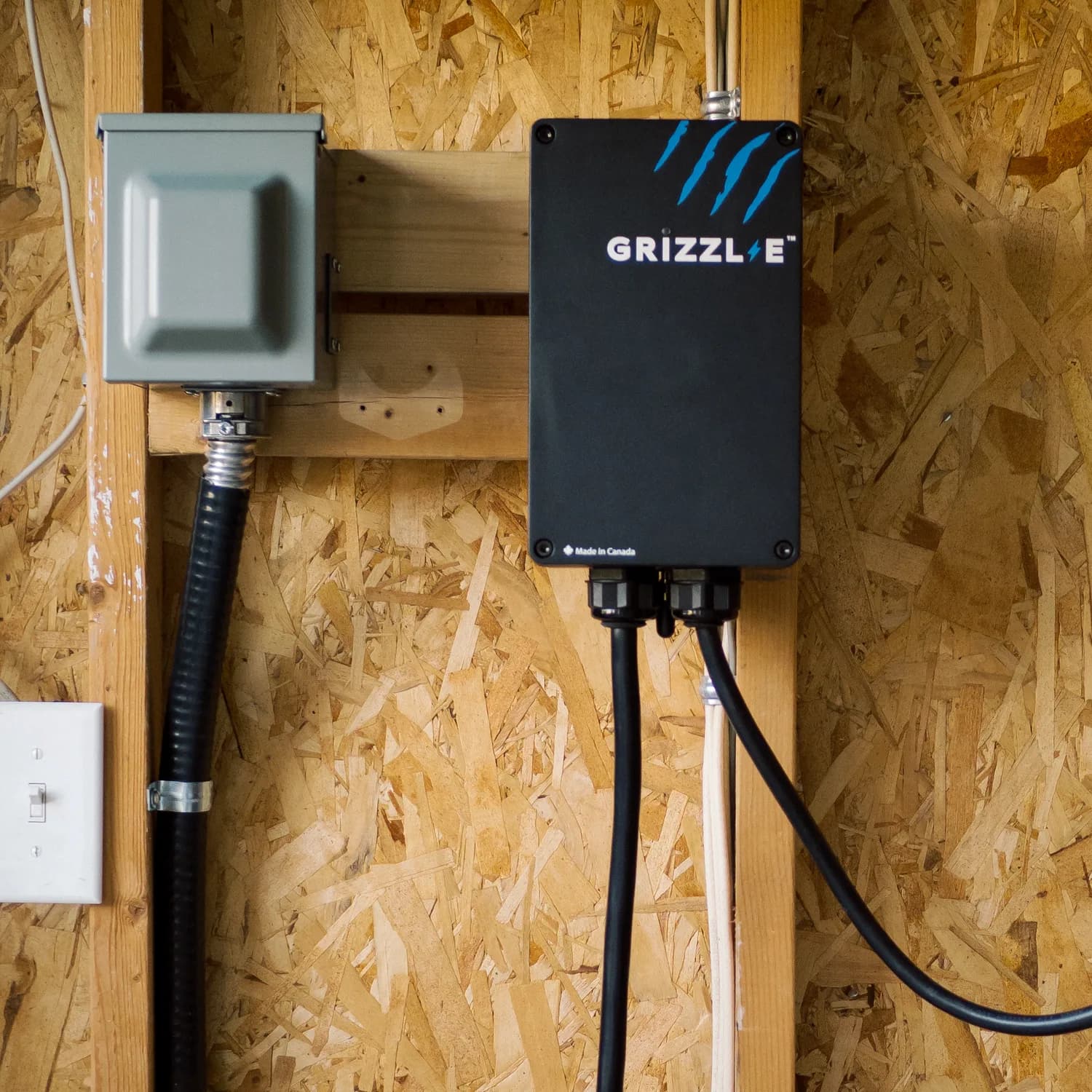A grey disconnect and a black Grizzl-E charger mounted side by side on a bare OSB garage wall.
