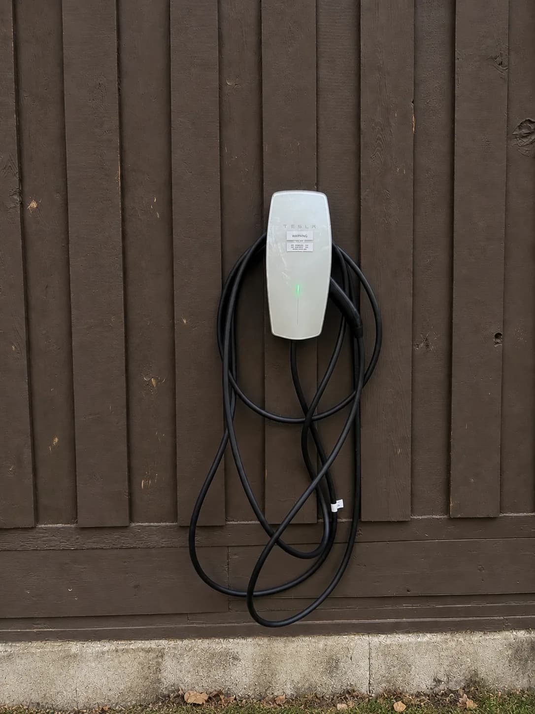 A Tesla Wall Connector mounted on dark vertical siding, its cable coiled beneath it.