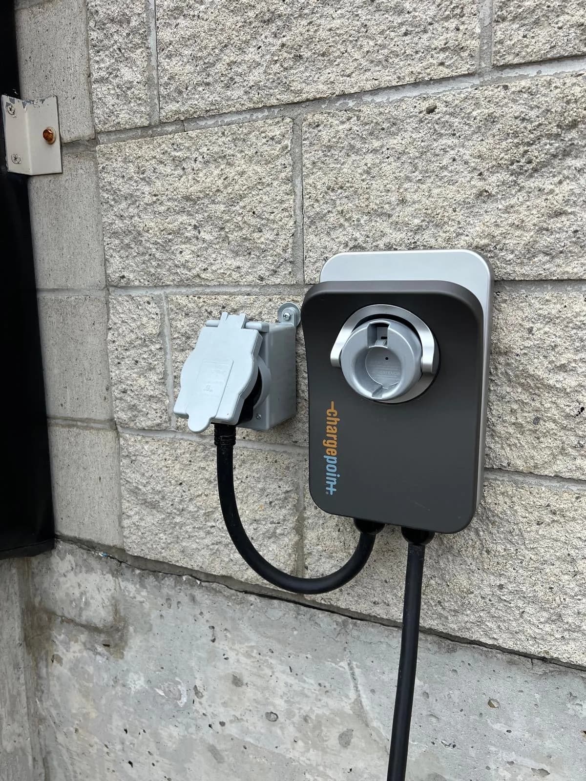 A ChargePoint charger mounted on a stone block wall beside a weatherproof outdoor receptacle, its cable running down the wall.