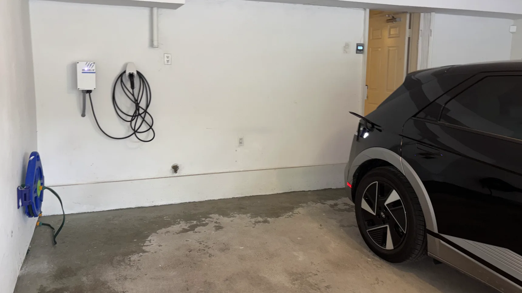 A Grizzl-E Avalanche Edition Level 2 charger mounted on a garage wall with its cable coiled on the holster and conduit dropped from the enclosure, the homeowner’s electric hatchback parked alongside.