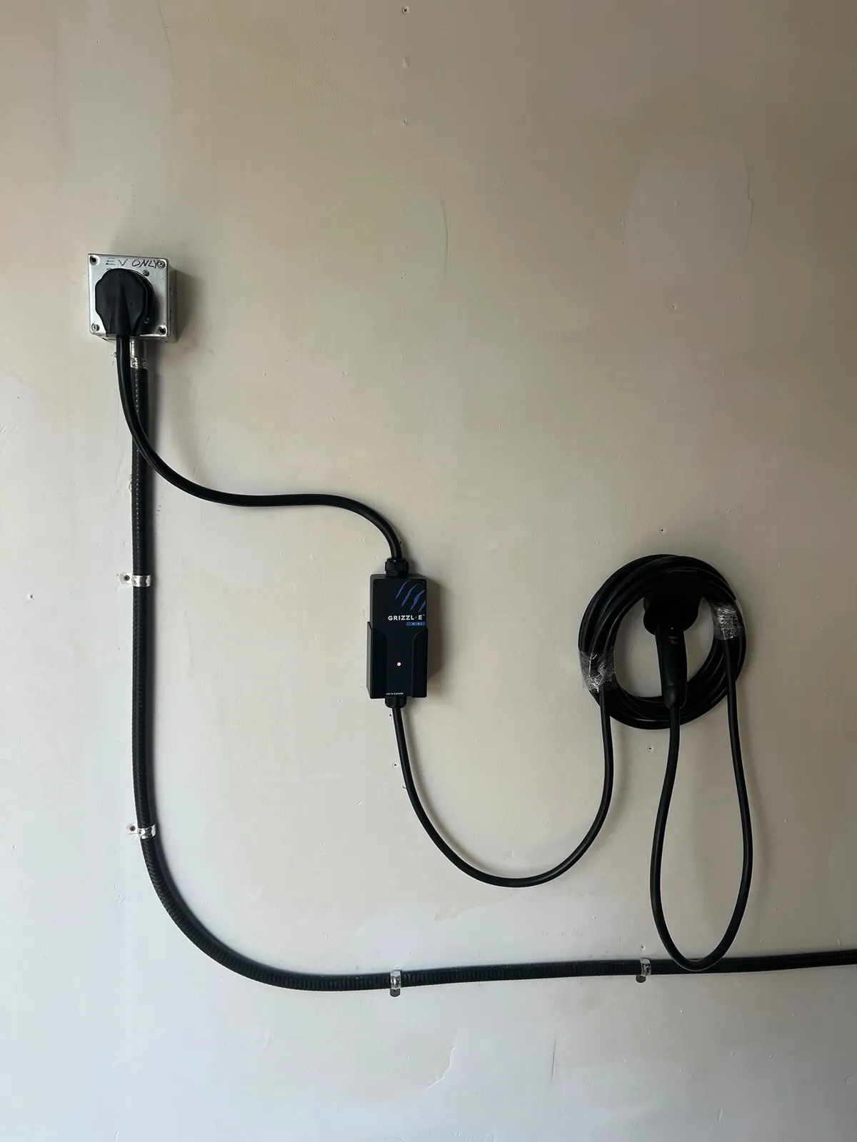 A Grizzl-E Mini charger on a pale wall, plugged into a metal-boxed receptacle marked “EV only”, with conduit clipped down and along the wall and the charging cable coiled on a hook beside it.