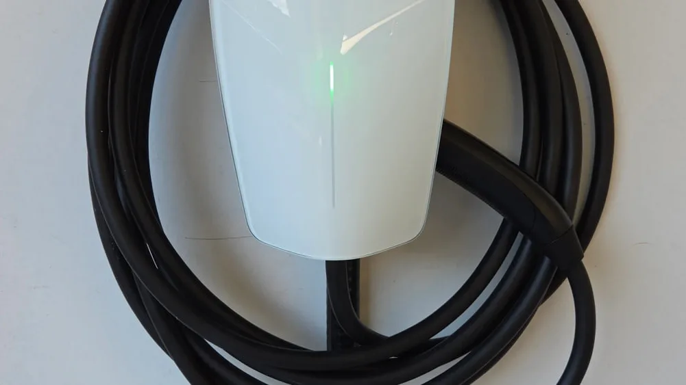 A white Tesla Universal Wall Connector on a pale wall, a green status light glowing down its face and its black cable wound in loops around it.