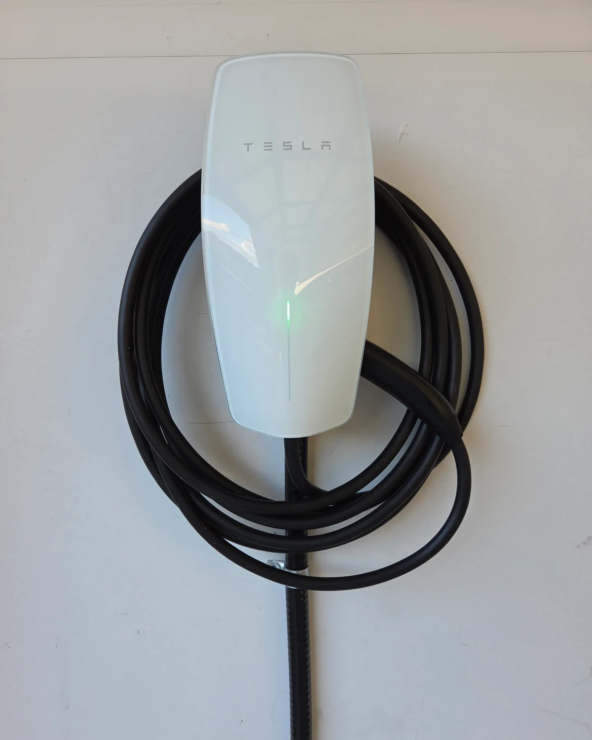 A white Tesla Universal Wall Connector on a pale wall, a green status light glowing down its face, its black cable wound in loops around it and the conduit running down the wall below.
